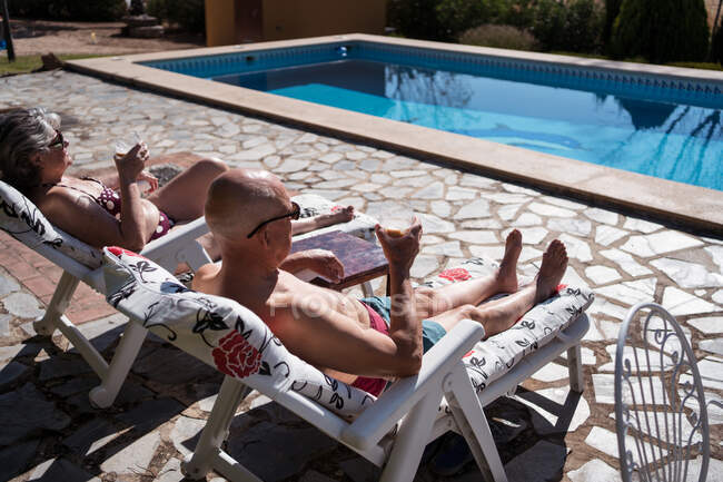 Вид сбоку на пожилую пару в купальниках, пьющую кофе со льдом во время отдыха на шезлонгах на заднем дворе — стоковое фото