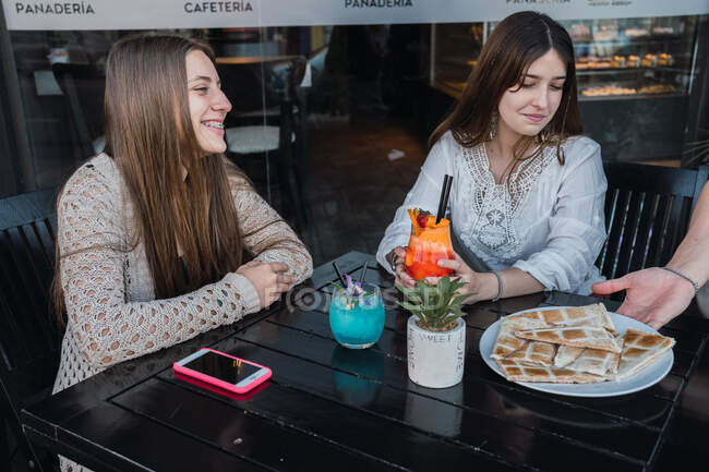 Crop content best female friends with tasty beverages while the waiter brings snacks in street cafeteria — Stock Photo
