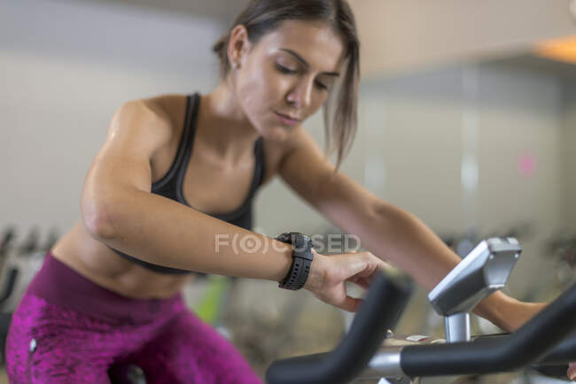Crop concentrated sportswoman exercising on stationary bicycle and checking result of cardio training on bracelet in gym — Stock Photo