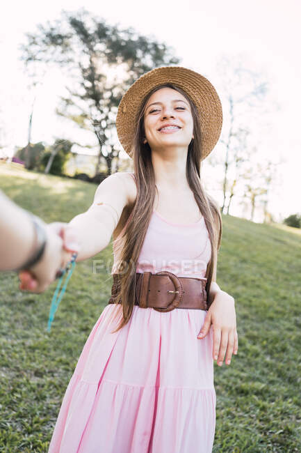 Усміхнена жінка-підліток у вечірньому та солом'яному капелюсі тримає анонімного партнера врожаю вручну, дивлячись на камеру в парку — стокове фото