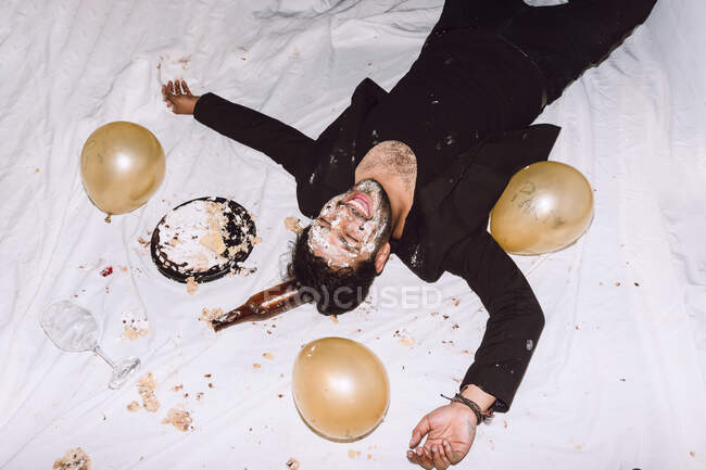 Bêbado rindo macho em bolo de aniversário esmagado deitado perto de garrafas vazias de cerveja e balões com os olhos fechados — Fotografia de Stock