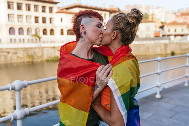 Cool mujer tatuada con mohawk y la bandera LGBTQ besando novia con los ojos cerrados contra el canal en la ciudad - foto de stock