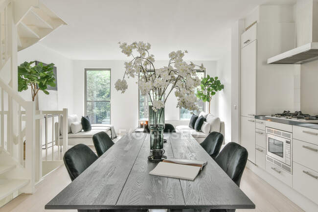 Interior de la zona de comedor con mesa grande con ramo de flores y sillas en apartamento moderno en el día y en el fondo la sala de estar y ventanas luminosas. - foto de stock