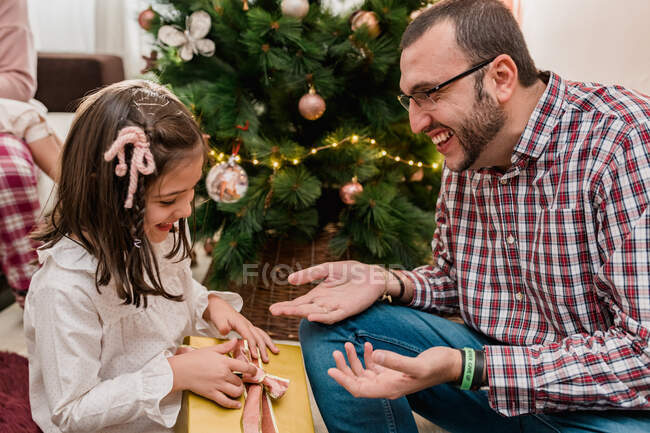 Alegre padre barbudo en anteojos sentado cerca del árbol de Navidad y dando caja de regalo a la chica feliz mientras celebra las vacaciones - foto de stock