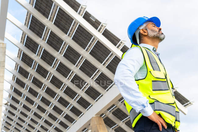 From below side view of mature ethnic male contractor in hardhat and vest breathing with closed eyes while standing near solar power station — Stock Photo
