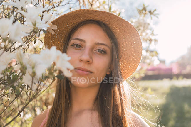 Charmante weibliche Jugendliche mit Strohhut und langen Haaren neben duftenden weißen Blume auf Strauch im Gegenlicht — Stockfoto