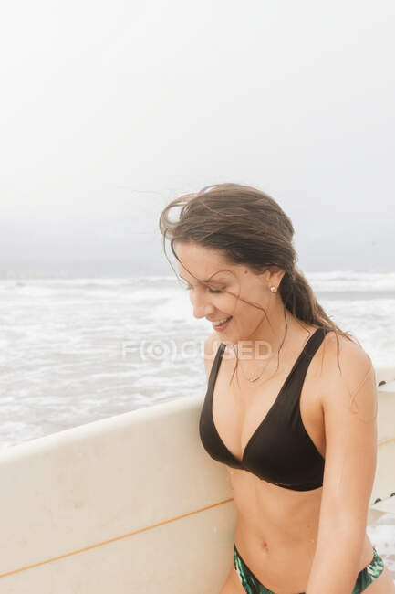 Вид сбоку на счастливую молодую спортсменку в купальниках с доской для серфинга, смотрящую на песчаное побережье против бурного океана — дневной свет, Море - Stock Photo