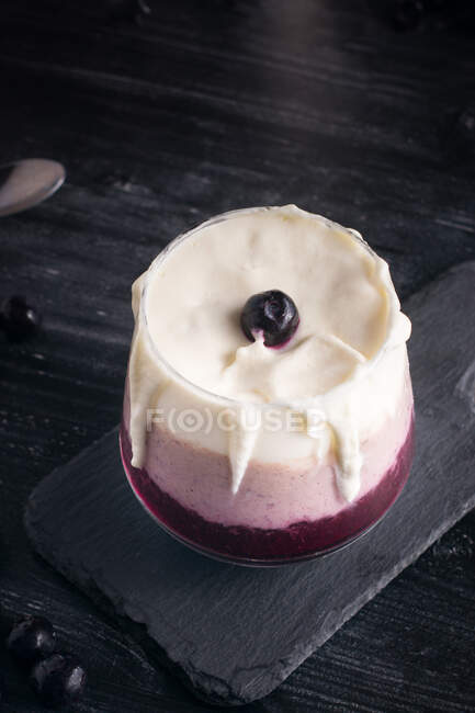 Glass of delicious banana and blueberry smoothie with whipped cream on chopping board against jar of yogurt and metal bucket — Stock Photo