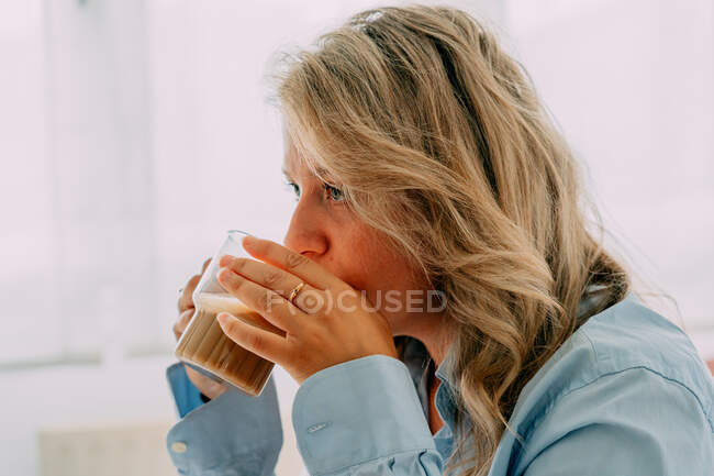 Vista lateral de la hembra adulta reflectante con cabello ondulado bebiendo sabroso café mientras mira hacia adelante en casa - foto de stock
