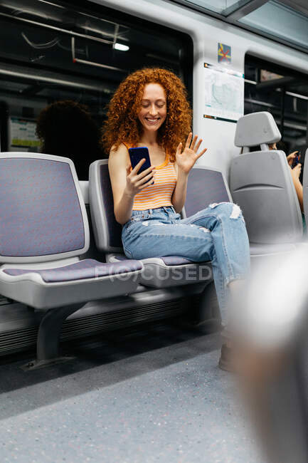 Дружелюбная женщина машет рукой во время видеочата по мобильному телефону, сидя со скрещенными ногами в поезде — стоковое фото