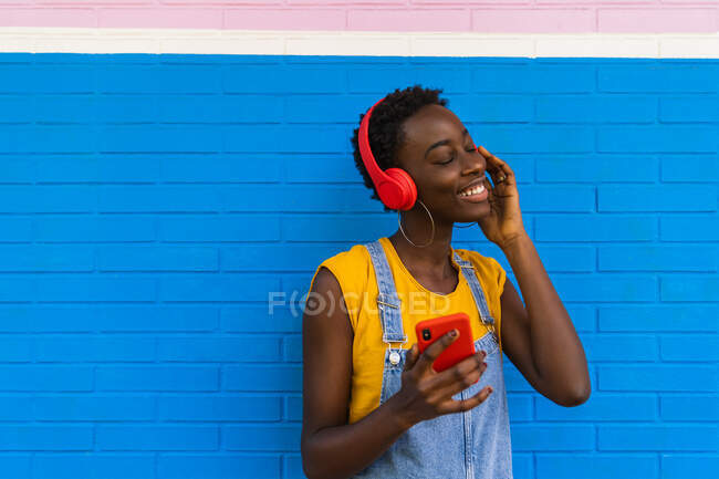 Encantada afro-americana fêmea desfrutando de músicas em fones de ouvido da lista de reprodução no smartphone contra a parede colorida — Fotografia de Stock