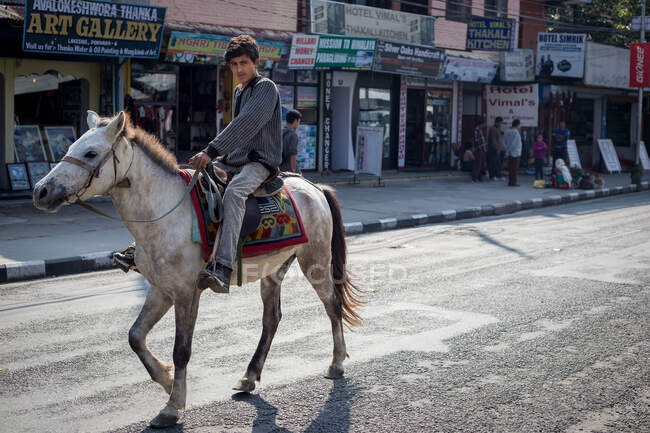 NEPAL, POKHARA - NOVEMBRO 6, 2015: Vista lateral da jovem fêmea em roupas casuais andando na estrada arenosa perto de barracas e capacete equitação cavalo no verão na cidade — Fotografia de Stock