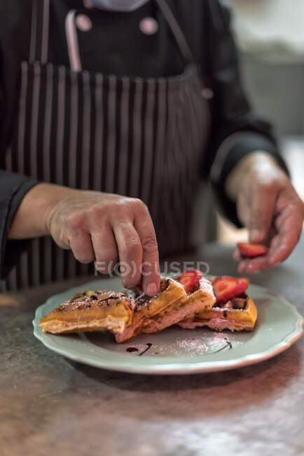 Crop anonymous male cook putting ripe strawberry slices on Viennese waffles with chocolate sauce and powdered sugar on plate in kitchen — Stock Photo