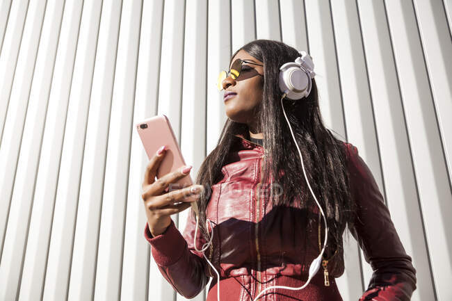 Низький кут нахилу молодих довговолосих афро-американських жінок у червоній куртці і сонцезахисних окулярах, які слухають музику через навушники і мобільний телефон, насолоджуючись сонячним днем у місті. — стокове фото