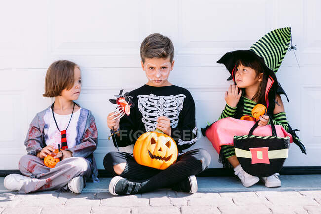 Full body of group of little kids dressed in various Halloween costumes with carved Jack O Lantern sitting near white wall on street — Stock Photo