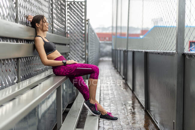 Vista lateral de cuerpo completo de atleta femenina tonificada en ropa deportiva sentada en el banco después del entrenamiento al aire libre - foto de stock