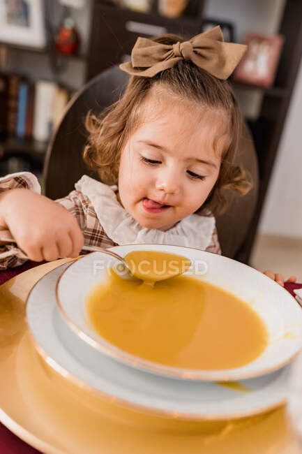 Charming child with bow on brown hair and spoon against plate of squash puree soup in house — Stock Photo
