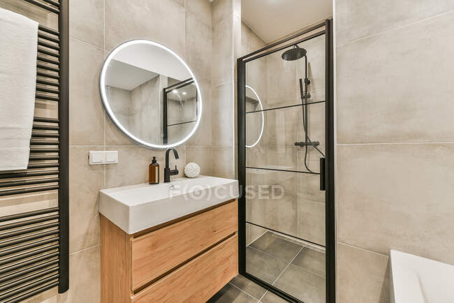 Banheiro empilhado com toalheiro aquecido perto de espelho brilhante pendurado acima da pia com sabão perto de cabine de chuveiro elegante e banheira — Fotografia de Stock