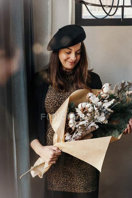 Femme positive dans des vêtements élégants et béret debout avec bouquet créatif avec des brindilles de coton et de sapin dans la salle de lumière — Photo de stock