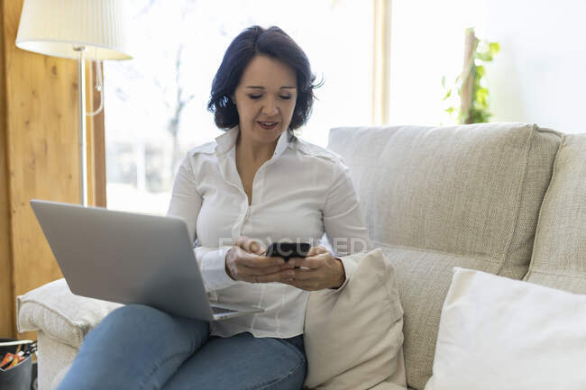 Feliz mujer madura freelancer navegando en el teléfono inteligente mientras trabaja en el ordenador portátil sentado en un sofá en casa - foto de stock