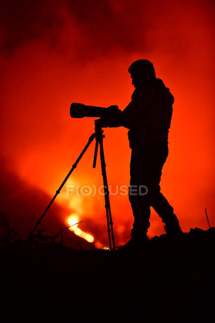 Vista laterale della silhouette di un uomo che registra e fotografa con un treppiede l'esplosione di lava sulle Isole Canarie La Palma 2021 — Foto stock