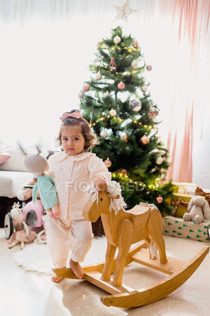 Чарівна маленька дівчинка, що стоїть біля дерев'яного коня під ялинкою, прикрашена казковими вогнями та іграшками — стокове фото