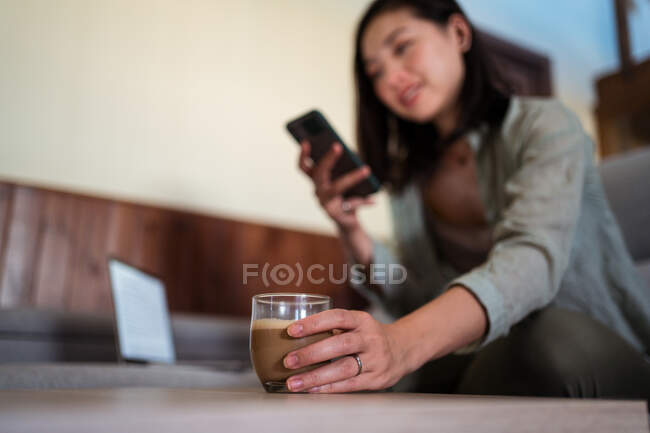 Jovem fêmea étnica com vidro de café surf internet no celular sentado no sofá na sala de casa perto do laptop — Fotografia de Stock