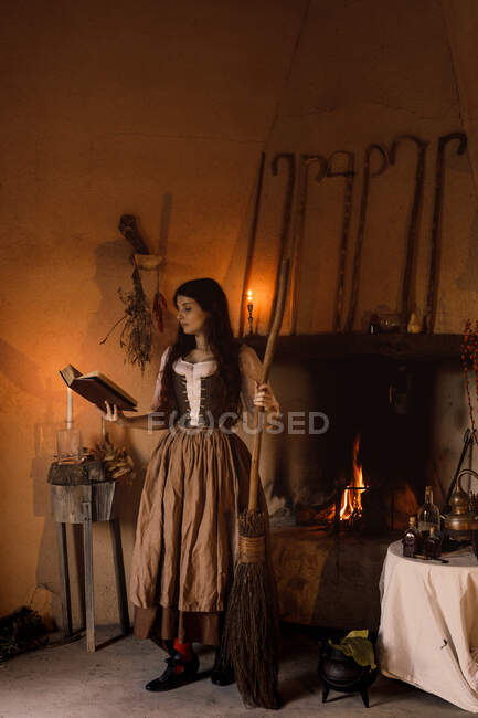 Hexe in langem Kleid liest Zauberbuch und steht in gemütlichem Raum mit Besen und Hexenkessel — Stockfoto