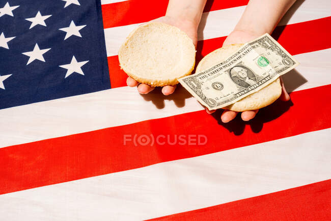 Desde arriba de la cosecha irreconocible persona con bollos mitades y billete de dólar sobre la bandera nacional americana en el Día de la Independencia - foto de stock