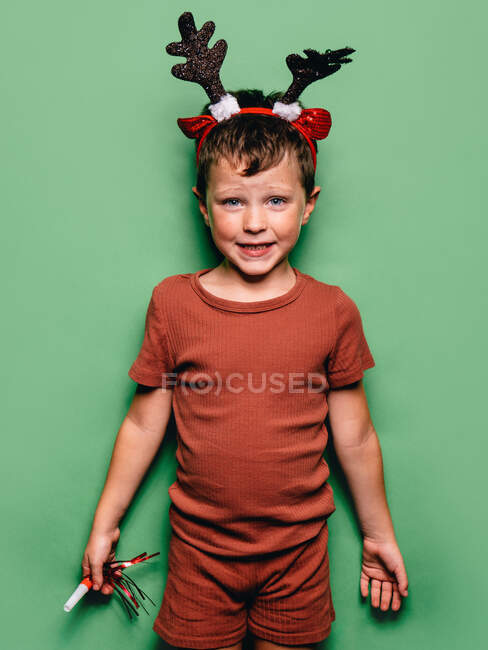Boy wearing reindeer horns headband and festive party blower standing against green background and looking at camera — Stock Photo