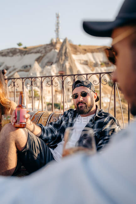 Homme barbu adulte dans des lunettes de soleil élégantes assis sur le canapé avec bouteille de bière près d'un ami de la culture en terrasse en Cappadoce, Turquie — Photo de stock