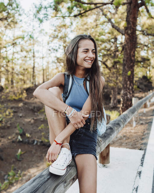 Adolescente sorridente na moda gumshoe tocando antebraço enquanto olha para longe na cerca em Tenerife Espanha — Fotografia de Stock