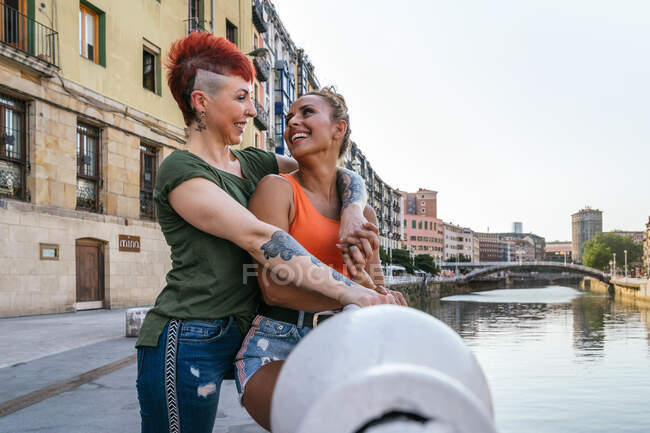 Side view of cheerful young homosexual woman embracing tattooed girlfriend with mohawk while looking at each other against canal in town — Stock Photo