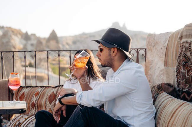 Vista laterale del giovane maschio barbuto in occhiali da sole e cappello elegante sorseggiando cocktail rinfrescante freddo mentre si rilassa sulla terrazza in Cappadocia, Turchia — Foto stock