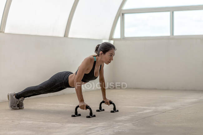 Side view of fit ethnic sportswoman in active wear exercising on push up bars while looking forward — Stock Photo