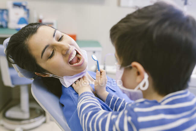Niño curioso en máscara médica jugando el papel de dentista y la comprobación de los dientes con espejo dental en el hospital - foto de stock