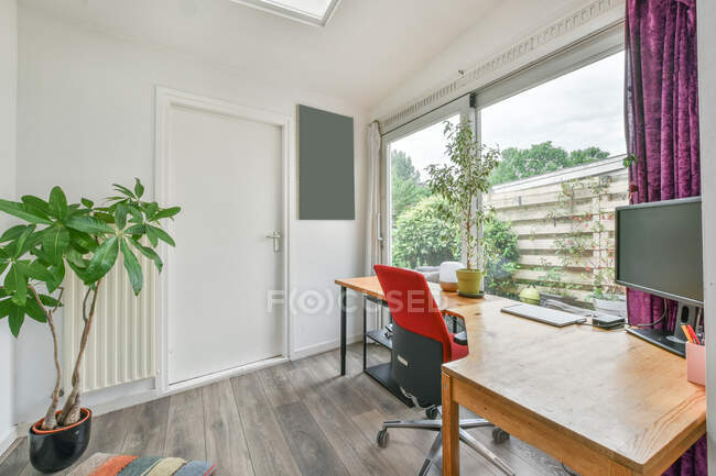 Scrivania da tavolo in legno con computer e piante posizionata vicino alla finestra in stanza luminosa in appartamento durante il giorno — Foto stock