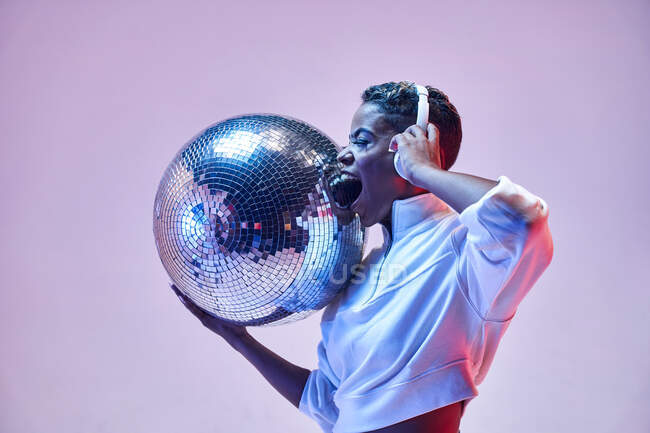 Coole ethnisch fröhliche Frau mit kurzen Haaren in drahtlosem Headset und trendiger Kleidung, die Hip-Hop mit offenem Mund und geschlossenen Augen tanzt und Glitzerball auf violettem Hintergrund hält — Stockfoto