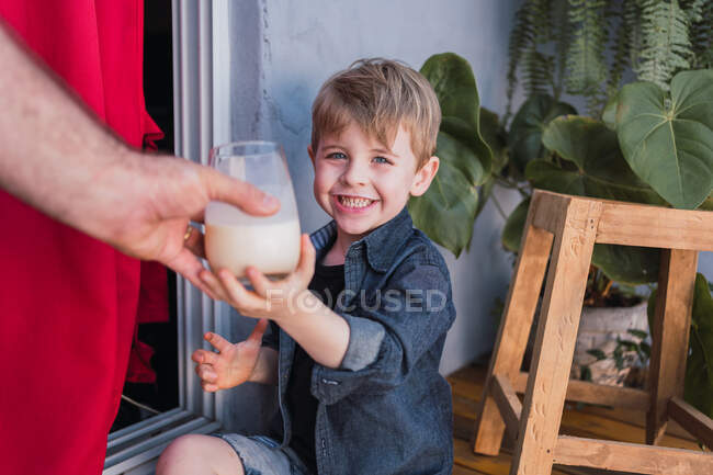 Alegre niño mirando a la cámara mientras recibe un vaso de bebida de la cosecha irreconocible papá contra el taburete hecho a mano - foto de stock