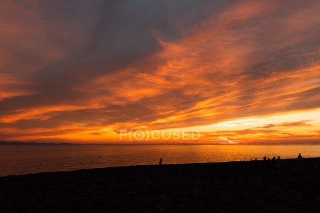 Vista espetacular das silhuetas de viajantes contemplando o oceano da praia sob o colorido céu nublado ao pôr-do-sol — Fotografia de Stock