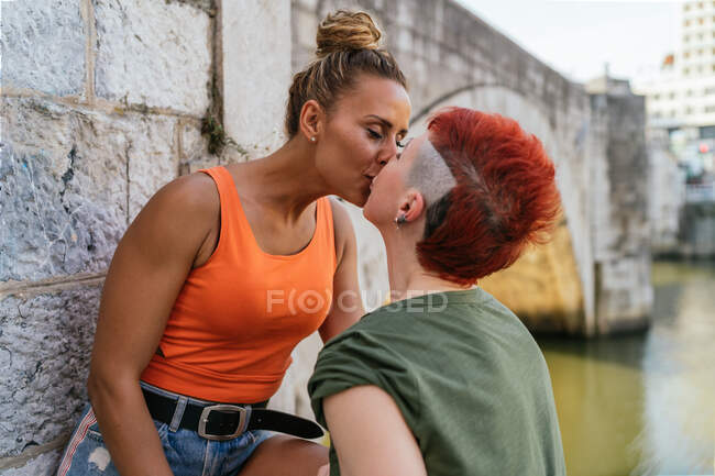 Vista lateral de feliz joven homosexual mujer con mohawk besos novia con los ojos cerrados contra el canal en la ciudad - foto de stock
