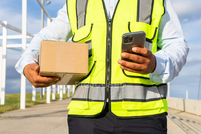 Ritagliato lavoratore maschile irriconoscibile in uniforme che trasporta pacco durante l'utilizzo del telefono cellulare al lavoro — Foto stock