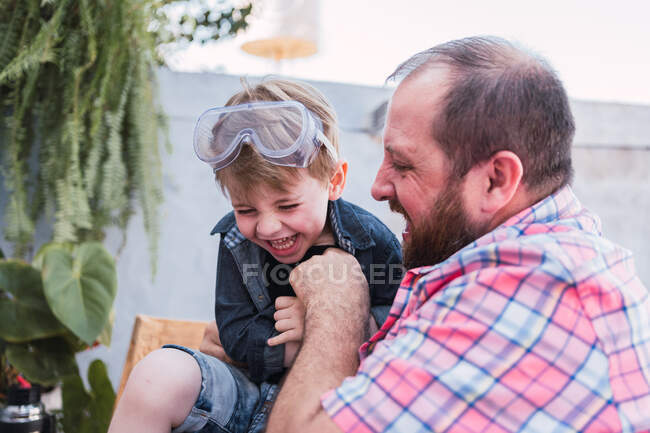Unrecognizable bearded dad in checkered shirt playing with cheerful boy in safety glasses while sitting in daytime — Stock Photo