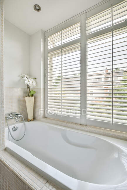 Белая ванна, установленная у окна в стильной ванной комнате с бежевыми плиточными стенами в современной квартире в дневное время — стоковое фото