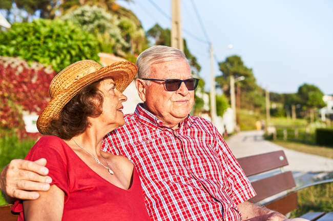 Side view of senior couple sitting looking away on wooden bench and enjoying summer day on shore of pond — Stock Photo
