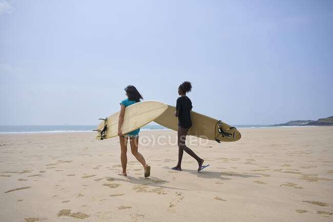 Vista posterior de jóvenes surfistas femeninas multiétnicas con tablas de surf hablando mientras pasean en la orilla arenosa y mirando y entre sí - foto de stock