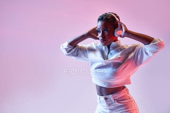 Mujer étnica enérgica en auriculares inalámbricos y ropa de moda bailando hip hop mirando hacia otro lado - foto de stock