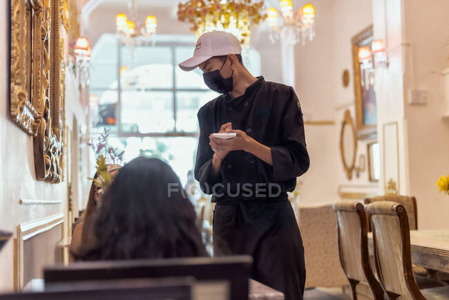 Dipendente caffetteria in uniforme e maschera di stoffa prendere appunti di ordine mentre si parla con donne irriconoscibili a tavola — Foto stock