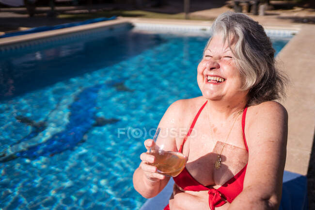 Hochaufragende Seniorin im Bikini lacht fröhlich, während sie am Pool mit Getränken chillt — Stockfoto