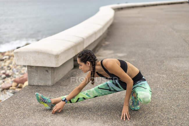 Seitenansicht einer fitten Läuferin, die die Beine streckt und sich beim Aufwärmen während des Trainings auf der Promenade auf eine Metallplanke stützt — Stockfoto